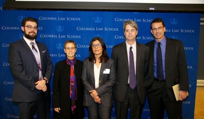 Five attendees stand together to take a picture. From left to right a gentleman in eyeglasses, dark facial hair stands in a charcoal suit, white shirt and multi colored tie.  Two women are both wearing eyeglasses dressed in dark pant suits.  The woman in the center wears a white blouse and badge clipped to her lapel.  The two men on the end are wearing eyeglasses dressed in dark suits. The gentleman to the farthest right wears a blue shirt and dark tie underneath.  