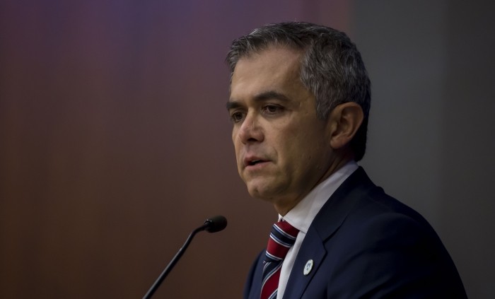 Profile angle of a man dressed in a dark blazer, white shirt and red, white and blue tie. He wears a tiny button on his lapel as he stands in front of a microphone awaiting to speak.