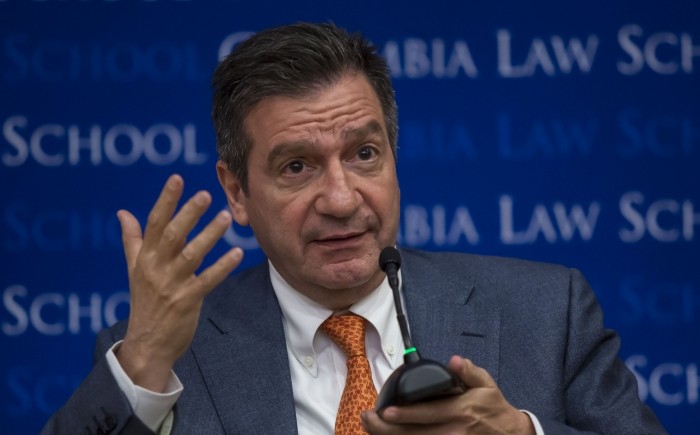 Man is engaged in discussion using a hand gesture while holding the base of a microphone with the other hand.  He wears a dark blazer, white shirt and rust colored tie.