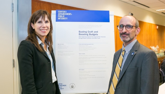 A woman and man are both dressed in dark blazers while a presentation board is positioned in the center of the two. They are smiling at the camera for the photograph.