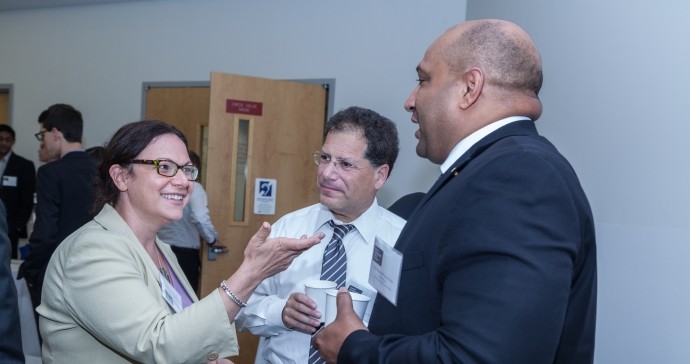 Woman is engaged in conversation with two gentlemen. She wears eyeglasses and a tan blazer. The gentlemen are dressed in buton down shirts, blazers and are holding styrofoam cups. 