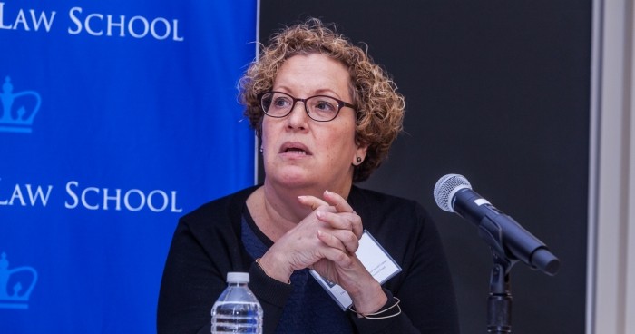 Woman wish short browin hair and dark rimmed eyeglasses sits while listening and clasping hands. She wears a dark long sleeved sweater.  A water bottle, reading materials and a microphone are positioned on the tabletop. 