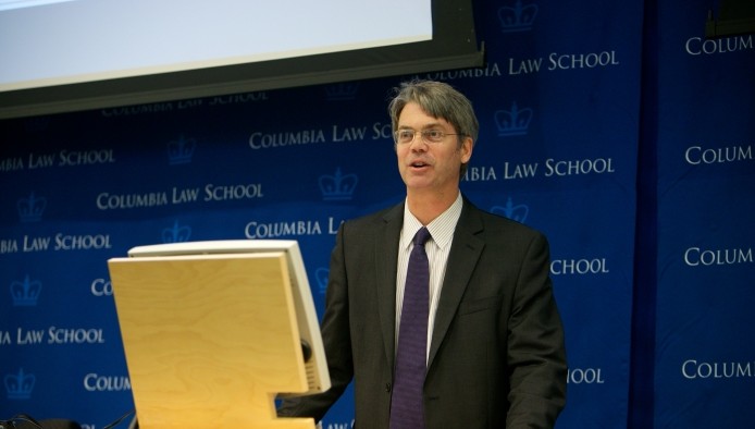 David Saltonstall is standing at a podium while wearing eyeglasses dressed in a dark blazer, white shirt and purple tie.   He is positioned in front of the royal blue printed Columbia Law School backdrop. 