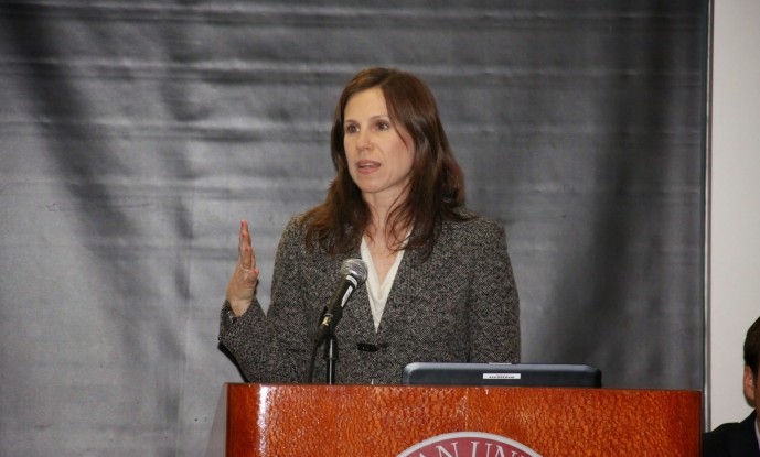 Jennifer Rodgers stands behind a podium wearing a kincaid blazer with a beige blouse underneath.  She gestures with her right hand.