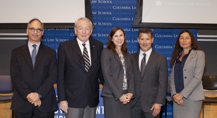 Group of five are dressed in professional attire while posing side by side for a photogaph.  In the backdrop are two overhead screens and the Columbia Law School background.