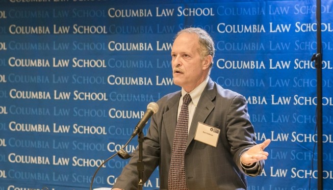 Man stands at podium dressed in a dark blazer, tie and white button down shirt.  He wears his ID badge.  He's gesturing with his left hand while standing behind a podium littered with papers.  He speaks into a microphone. The Columbia Law School backdrop hangs in the rear.