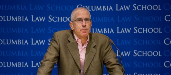 Man wearing eyeglasses with an olive colored blazer and pink button down shirt holds the podium while staring straight ahead.  The Columbia Law School backdrop is in the rear.  A microphone, bottled water and papers are on top of the podium.