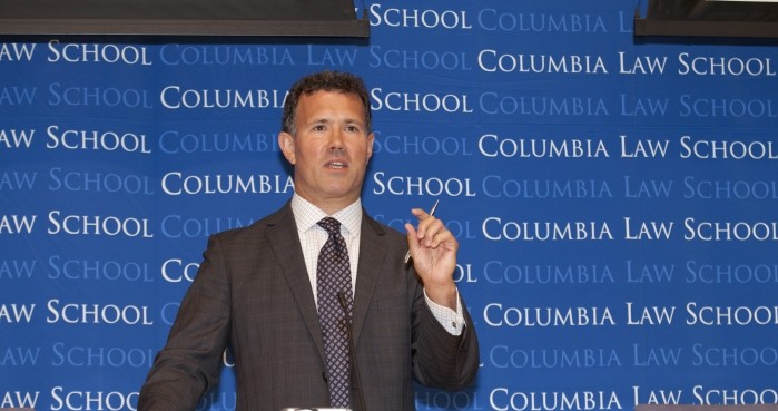 Man speaks at a podium stage holds pen in left hand. He's wearing a dark suit with a white buttn down dark tie detailing a button on the upper left lapel.  The Columbia Law School backdrop falls in the background. 