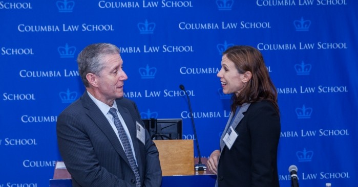 In the far background is the royal blue Columbia Law School imprinted backdrop.  A man and woman are engaged in conversation facing each other both are dressed in dark blazers and are smiling. 