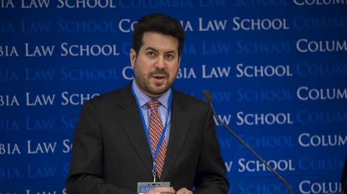 Man stands behind podium wears a dark blazer, light blue button down shirt and red striped tie. An ID badge hangs from his neck.  His hands are closely positioned on top of podium and atop of a booklet.  The Columbia Law School backdrop is on the rear wall.