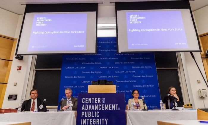 Panel of four are seated at a long table. There are two overhead screens hanging above with a curtain backdrop of the Columbia Law School imprint.  There is a podium in the center stage displaying a covering displaying the words Center for the Advancement of Public Integrity