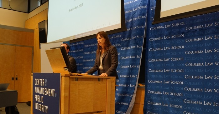 A woman stands at a platform viewing a monitor while two screens are hanging behind her along with a backdrop curtain with Columbia Law School logo.