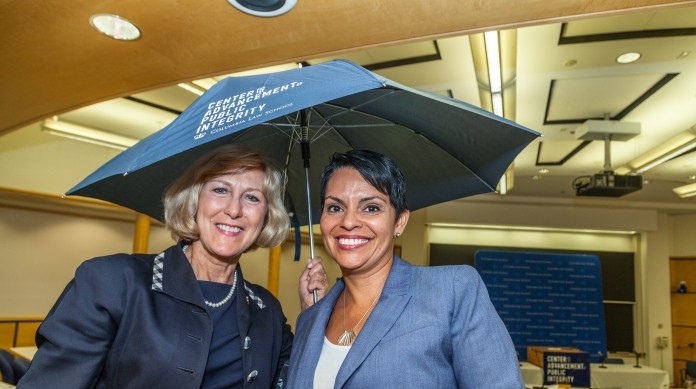 Two women are dressed professionally while holding an umbrella and smiling for the camera.