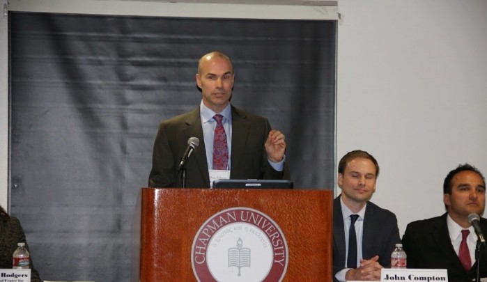 Douglas Wilmore is standing behind a podium. He wears a dark blazer, light blue shirt and maroon tie.  He gestures with his left hand. There are two gentlemen seated at a table covered with a white tablecloth. The panel of men each one wears a dark blazer with a name card, water bottle and microphone positioned the table. 