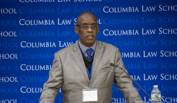 Man stands behind a podium with a Columbia Law School backdrop positioned in the backdrop.  He wears a light grey blazer, darker vneck sweater underneath with a light blue colored shirt and royal blue tie.  He wears eyeglasses and a plastic ID badge around his neck.  There is a bottle of water and microphone on the podium as he speaks.