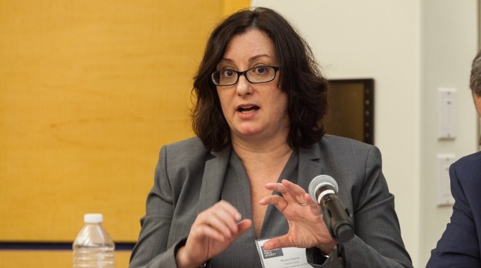 Woman with dark hair and eyeglasses wears a dark vneck blouse and dark suit with ID badge at a panel.  She is using her hand gestures during speech. A water bottle sits on the tabletop . 